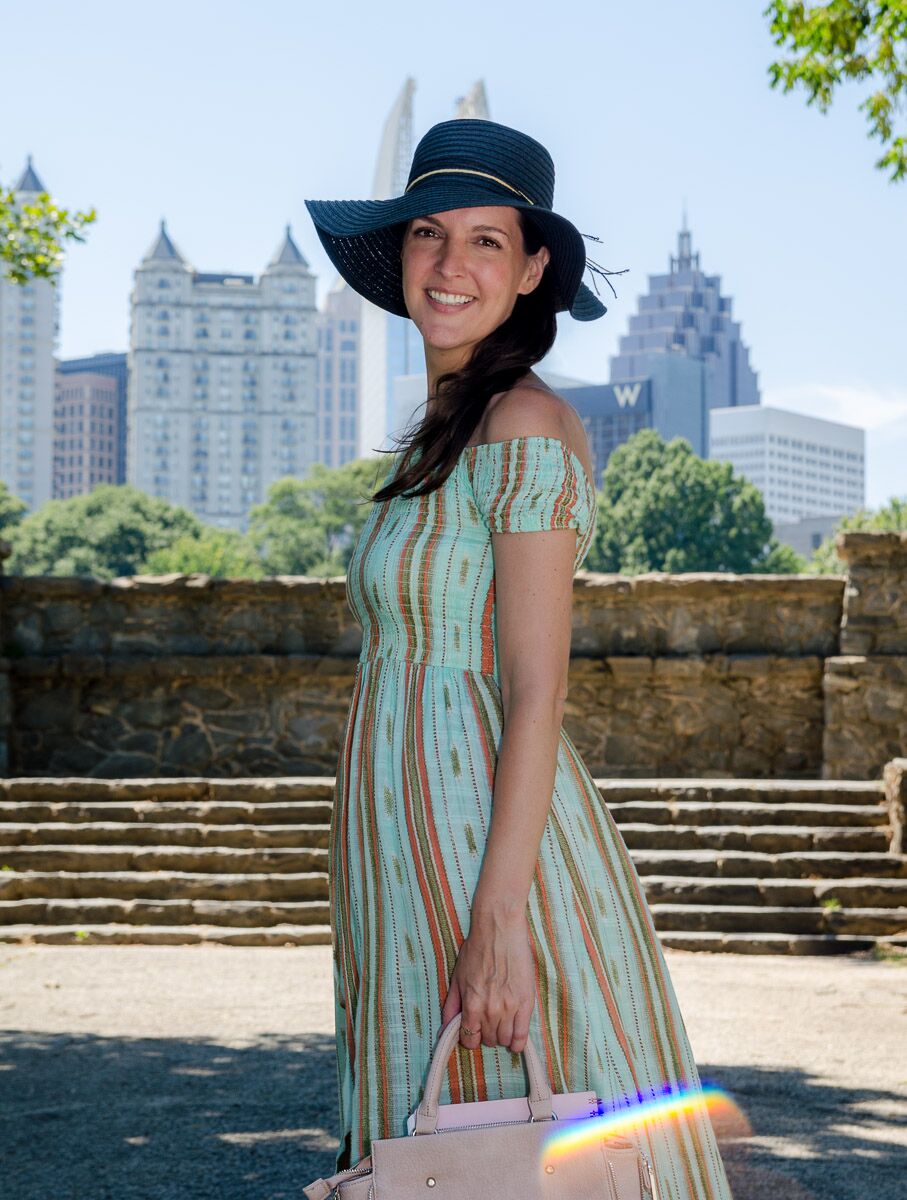Amy Westmoreland, manifesting teacher, smiling in a light blue and orange striped sun dress and a wide-brimmed black hat. She stands in a sunlit park with a city skyline in the background. A small, vibrant rainbow light flare appears across the bottom of the image.