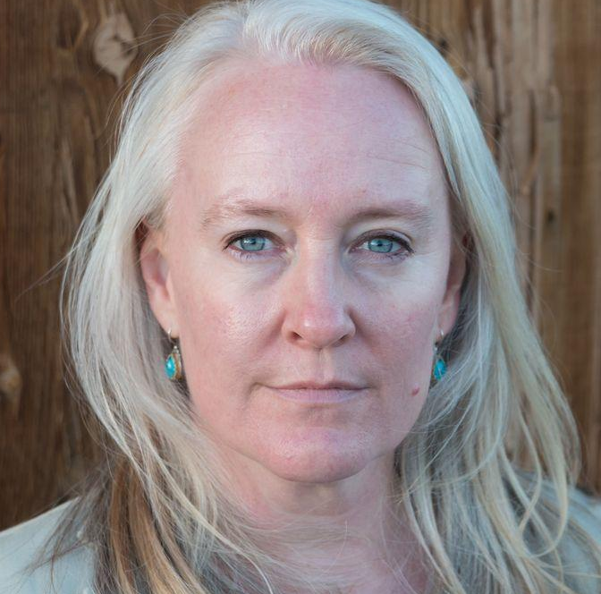 A professional headshot featuring Bridget Thomas, a coach and author, with blonde hair and blue eyes, smiling directly at the camera against a rustic, dark wood panel background. She is wearing turquoise teardrop earrings.