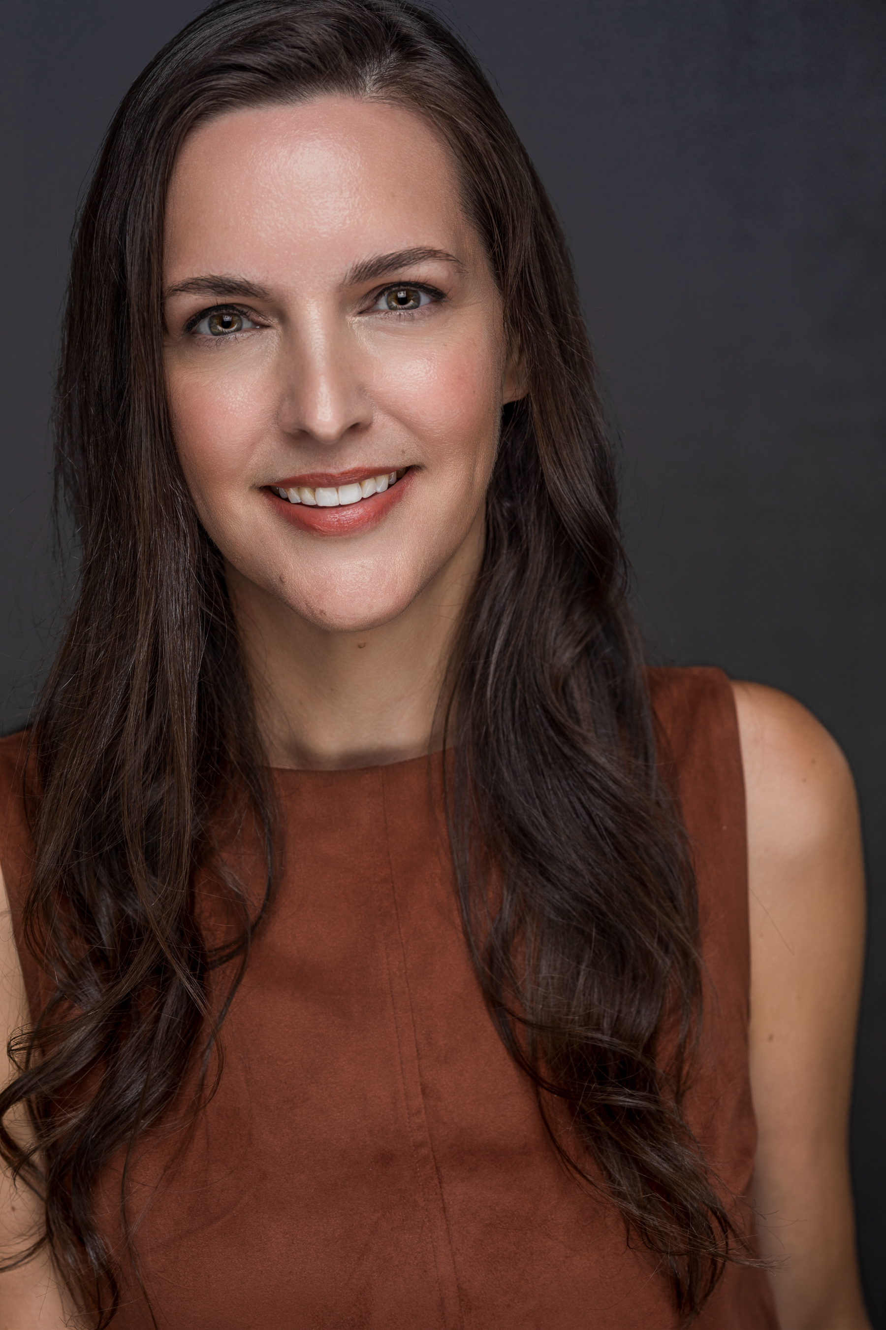 Amy Westmoreland, founder of Illuminating Joy, smiling in a professional headshot. She has long, dark hair and is wearing a rust-colored sleeveless top against a neutral dark background.