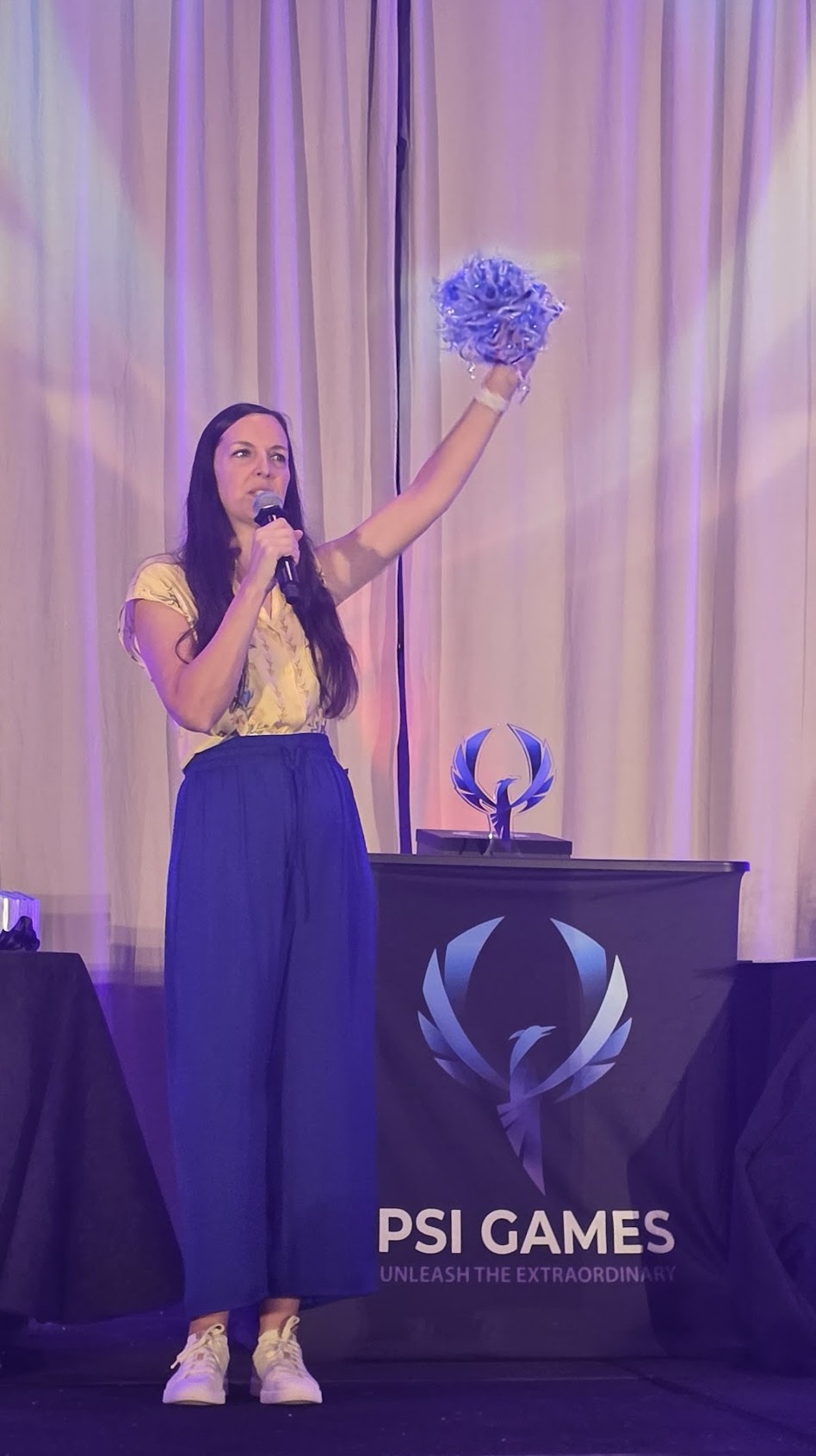 A full-length photograph of Amy hosting on stage at Psi Games 2026. She is wearing a yellow patterned top and flowing cobalt blue pants, and she is speaking into a handheld microphone with her left arm. She is holding up a blue crystal award for the crowd to see. Behind her is a dark podium with a blue phoenix-style trophy on top and a banner featuring the 'Psi Games' logo and the slogan 'Unleash the Extraordinary.' The background features white curtains illuminated with purple light.
